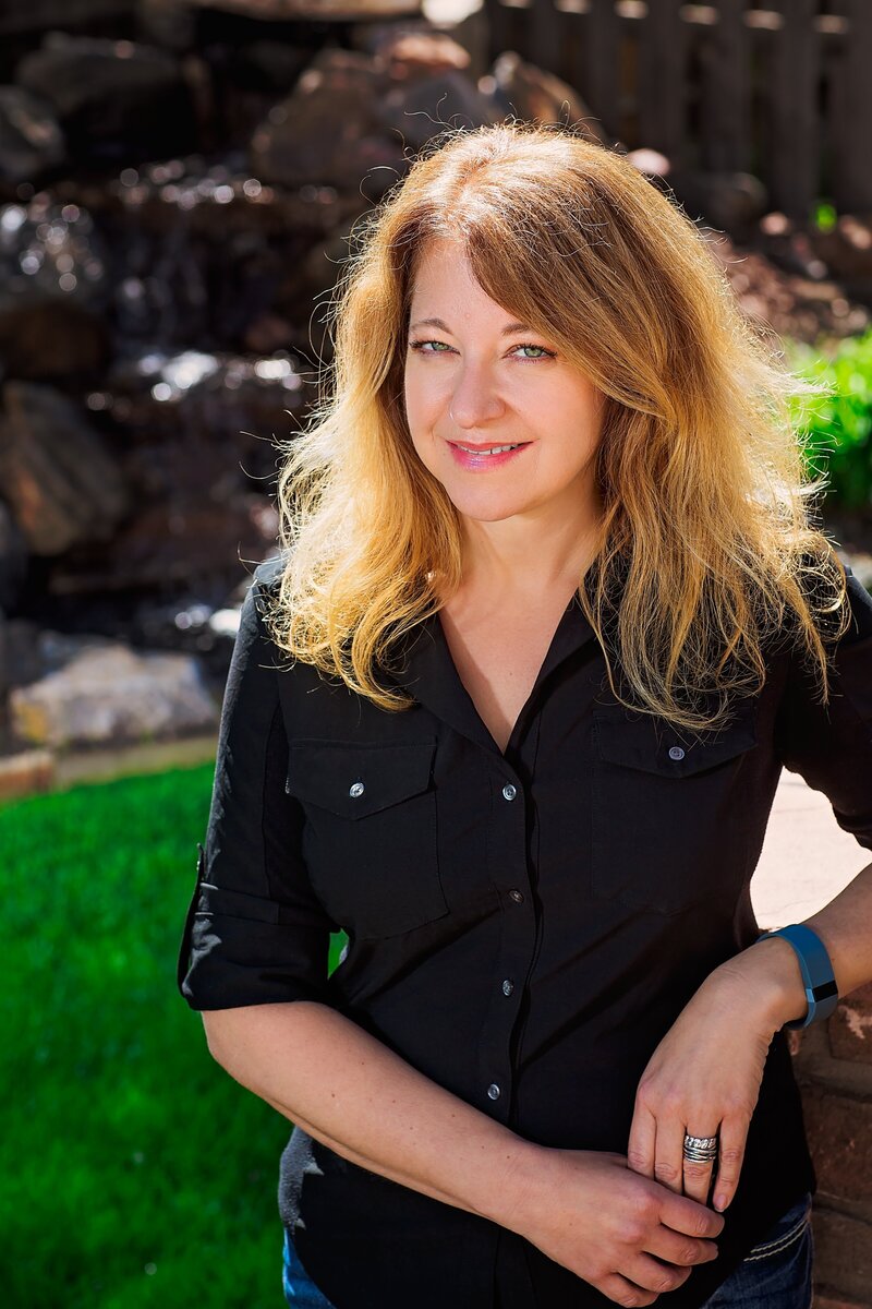 Cyndi Stewart poses outdoors in bright sunshine, leaning on a rock wall with a gentle smile.