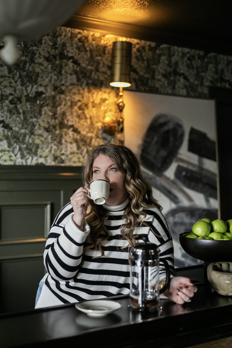Ashleigh Henry, life coach based in Asheville, North Carolina is sipping coffee from a french press in The Blind Tiger, near The Biltmore House and Riverarts Distrct, with green floral wall paper and a black and white painting in the background, she, too, is wearing a black and white sweater