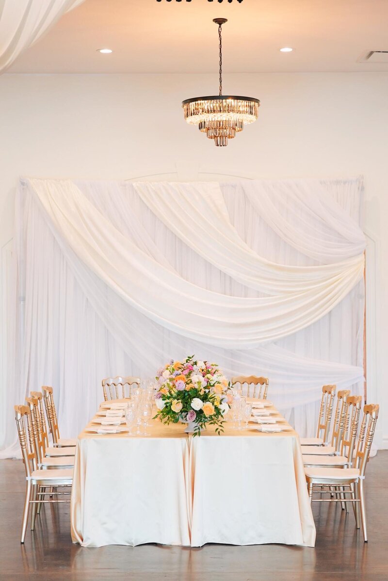 A decorated wedding table with gold chairs, a floral centerpiece, and white draped fabric backdrop under a chandelier,  at Grbic Events, a wedding venue in St. Louis.