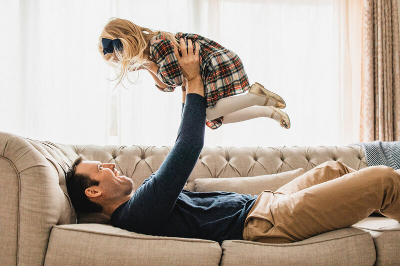 Father playing airplane with daughter on white couch in front of a big  window