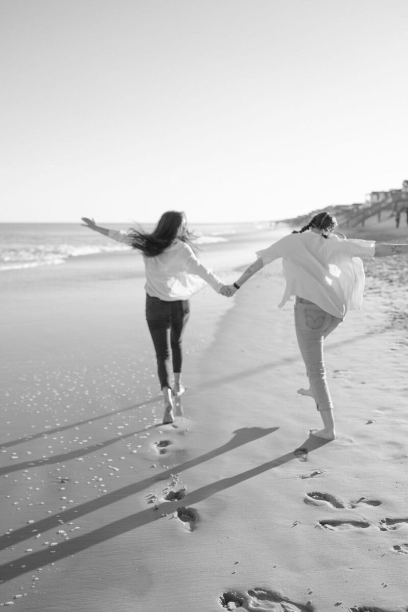 two friends hold hands and run barefoot down the beach towards the waves