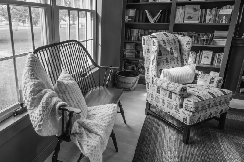Black and white photo of library with antique spindle bench with cream knit blanket, early american textile covered wingback chair with built-in bookshelves with books on the shelves