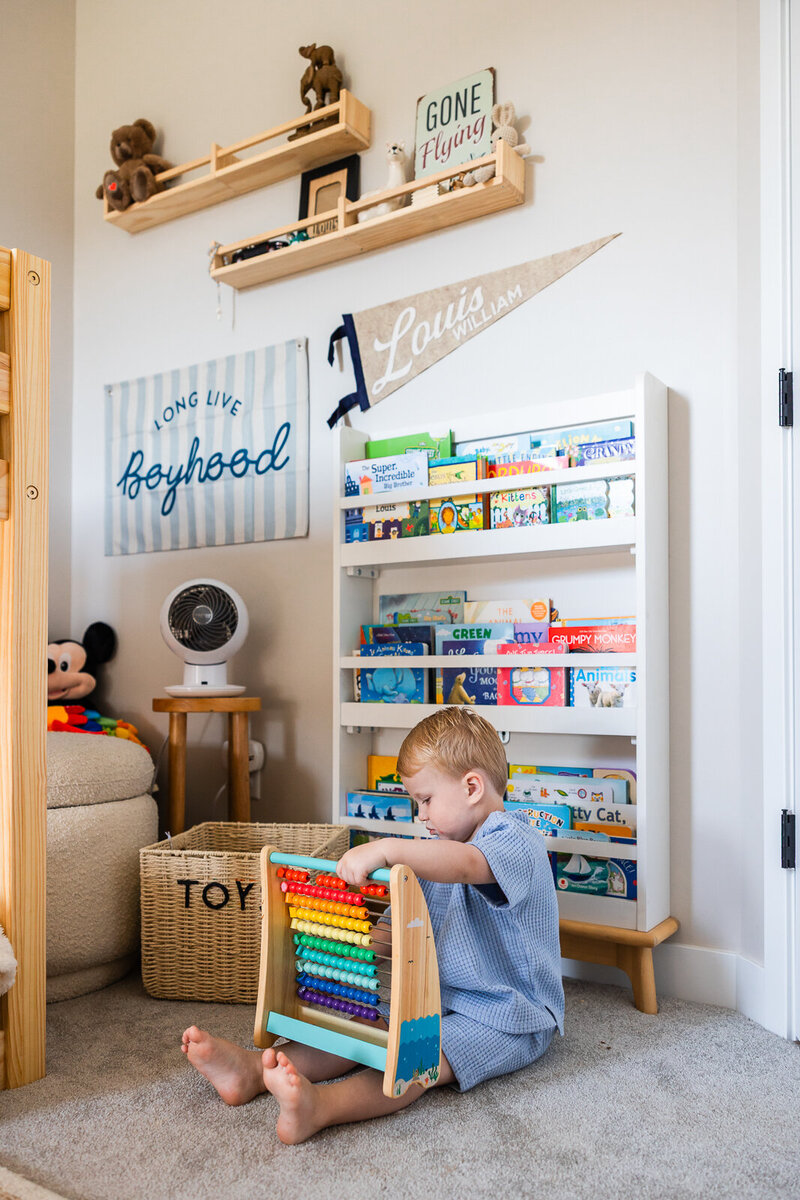 Toddler boy sitting on his bedroom floor next to his bookshelf and toy box playing with a toy made of beads. 