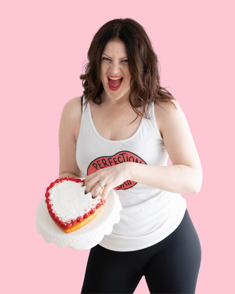 Emily, smiling with crumbs around her mouth, wearing a white tank top, holding a heart shaped cake with one hand smashing the icing.