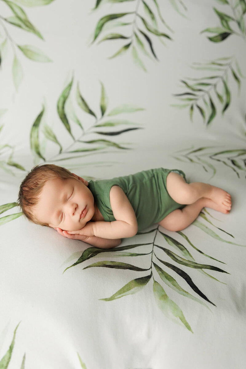 baby boy sleeping with his hands under his head in a green onesie on a green plant background newborn harrisburg photos