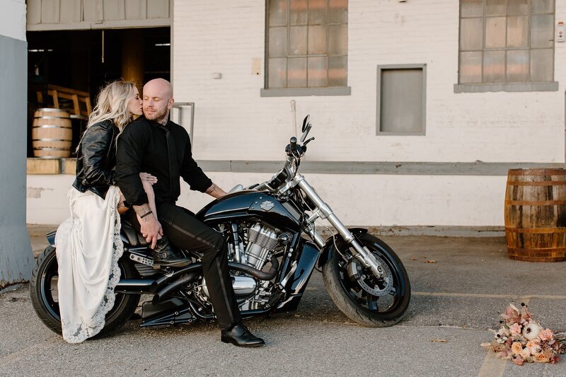 A wedding couple sitting on a motorcycle while one kisses the other's cheek 