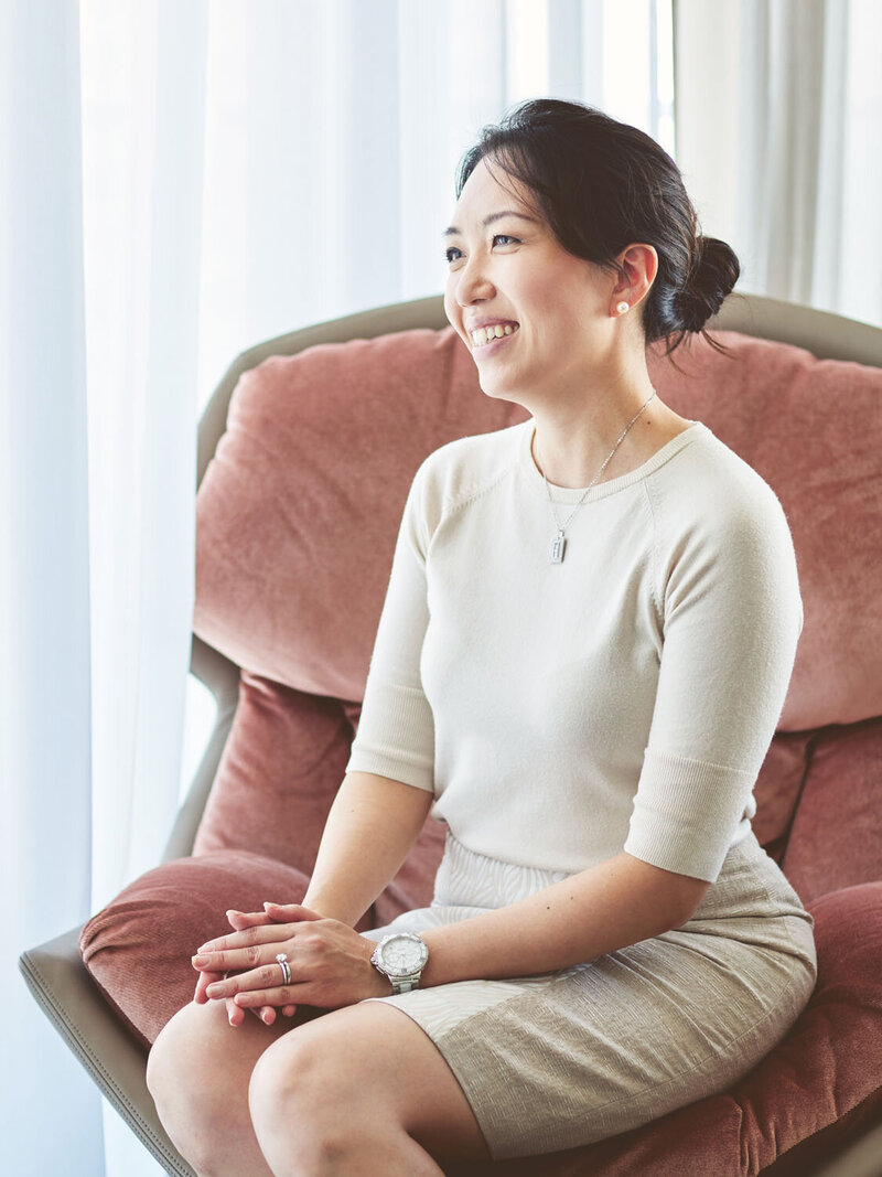 Christen elegantly dressed sitting in a designer arm chair by a big window with sheer curtains