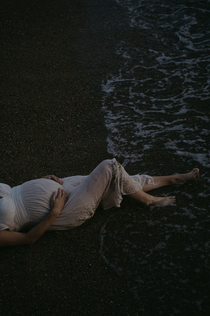 Femme en robe blanche et ventre rond allongée sur le sable noir à la tombée de la nuit. Séance photo grossesse vers Nantes en Loire Atlantique.