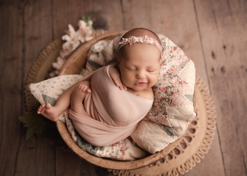 Baby girl is wrapped in pink stretch fabric with her legs out and folded gently atop of her. She is placed in a wooden bowl atop of a folded vintage quilt. The baby is sleeping peacefully. Captured by best Temecula Newborn Photographer Bonny Lynn Photography