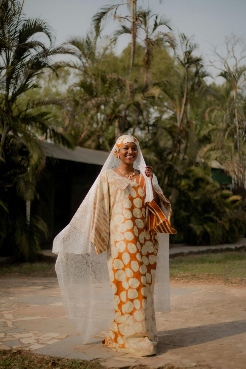 Smiling African bride in traditional patterned attire and lace veil walking outdoors, captured in natural light by Chi Ramos Photography — multicultural wedding photographer.