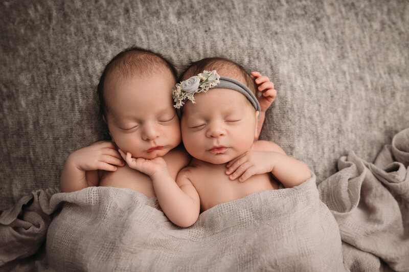 Newborn boy girl twins laying down tucked in a grey blanket holding each other side by side