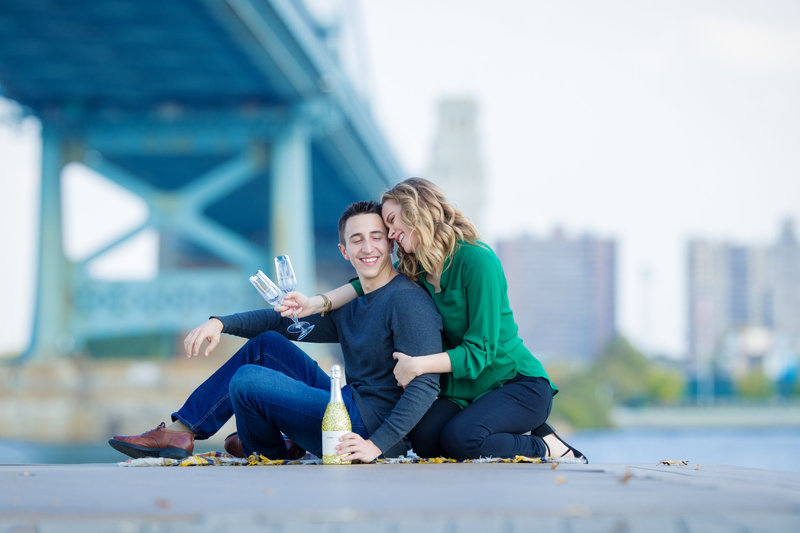 Engaged couple sharing a celebratory moment with champagne on Race Street Pier, surrounded by stunning city views.