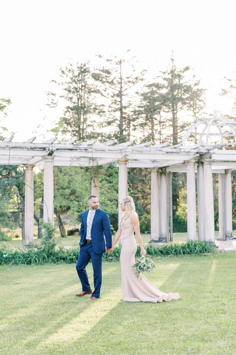 Couple walk hand in hand through a field with marble architecture.