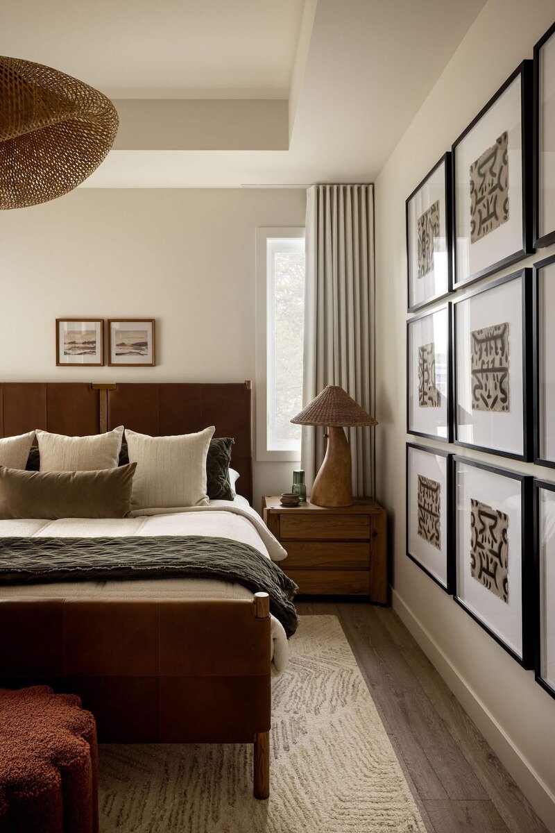 Japandi-style primary bedroom with leather king bed, rattan pendant light, tribal abstract artwork, boucle ottomans, and layered bedding.