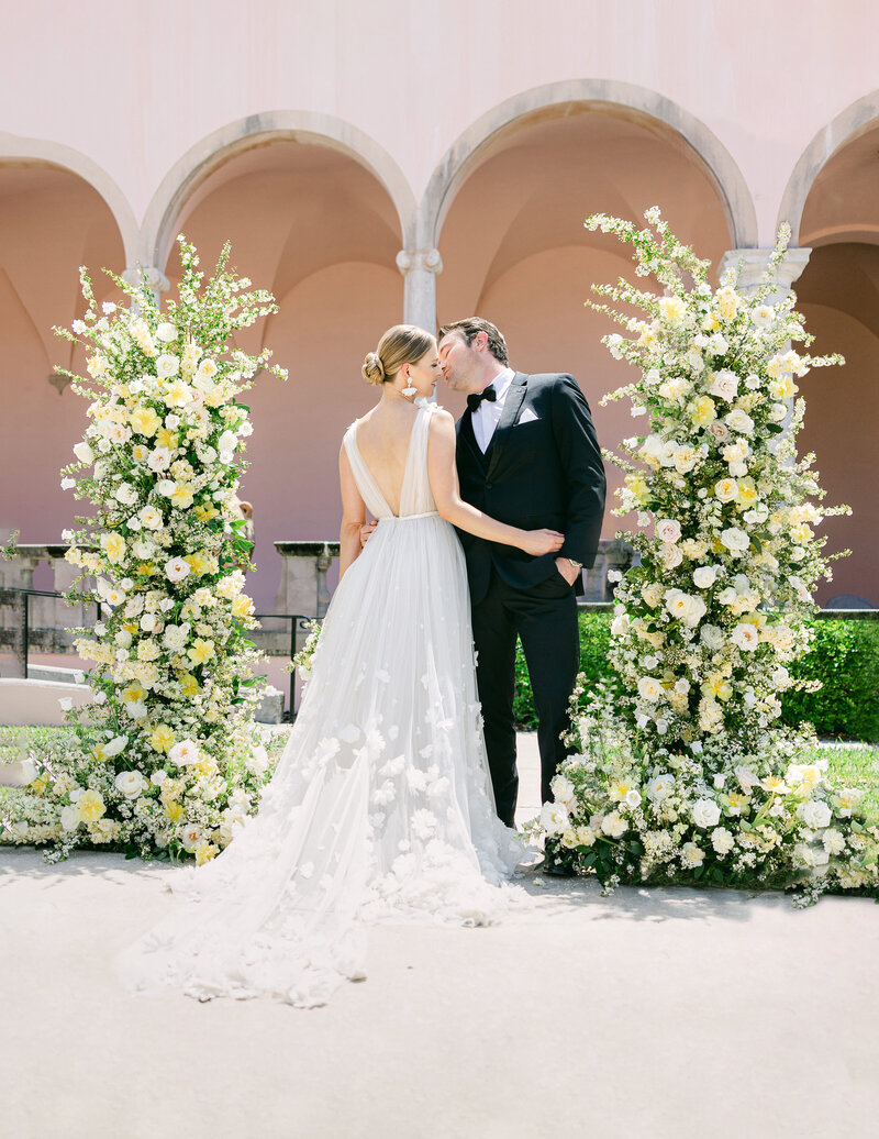 A bride and groom share a romantic moment beneath the arches of The Ringling Museum in Sarasota, surrounded by lush floral arrangements, captured by Amia Marcell destination wedding Photographer