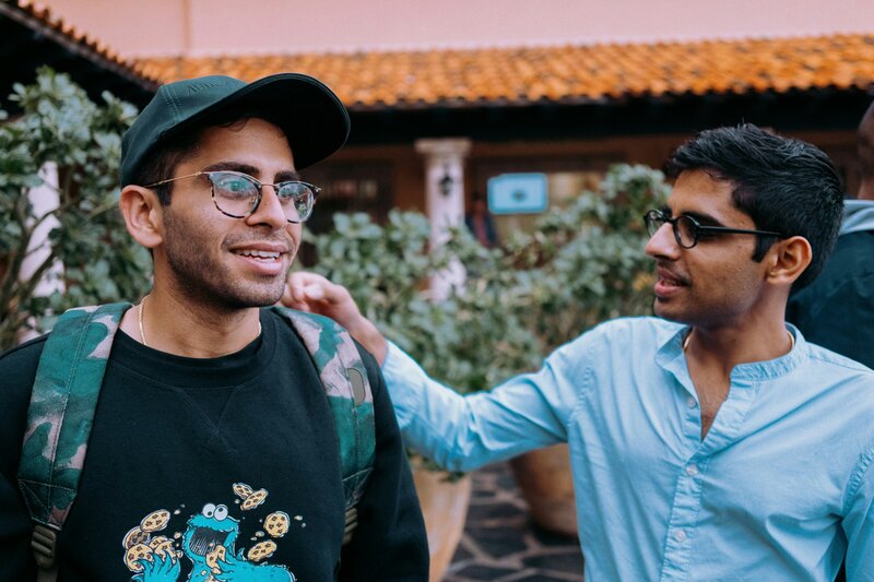 Two young men, one wearing a backpack and baseball cap, interacting in a courtyard, illustrating friendship and connection.