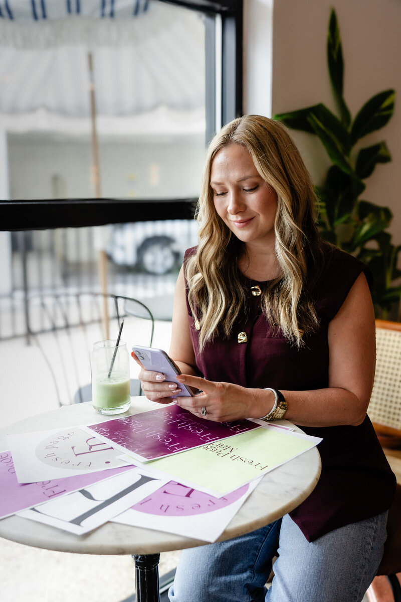 Woman using phone at a cafe with Fresh Page Marketing brand sheets, representing Shopify store management, Faire wholesale growth, and website strategy.