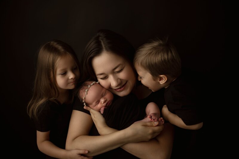 Mother holding newborn with two siblings smiling close - family newborn photography in Maple Grove Mn studio