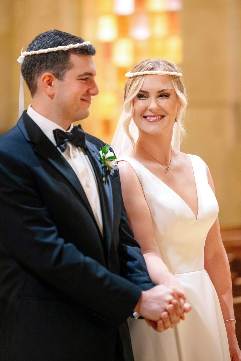 A bride and groom stand together during a wedding ceremony inside a church. They both wear white headbands and hold hands, smiling at each other. She is in a white gown and veil, and he is in a black tuxedo with a bow tie.
