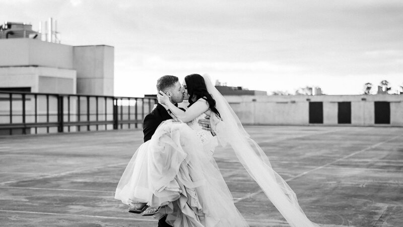 Groom carries bride in his arms as they kiss on a rooftop