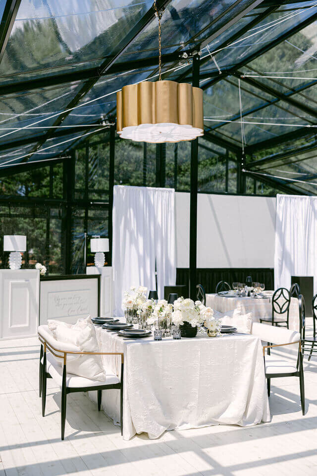 A glass tent showcases a white table adorned with refined black and gold accents, providing wedding inspiration and elegant decoration.
