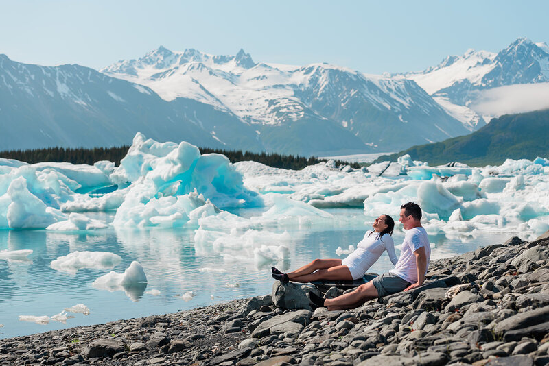 Filling the background is innumerable blue-tinted white icebergs floating in the beautiful turquoise water of Bear Lake during a two day luxury elopement with dogsledding in Alaska.  Behind that are rugged snow capped mountains On the right side in the front of the the frame, the bride and groom lounge in the sun on large flat stones on the rocky shoreline --they are both wearing white swimwear, and have just done a cold plunge during their elopement.