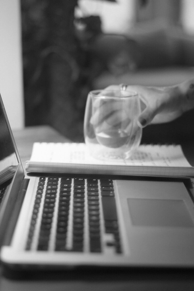 A side view of a work desk captures an open laptop and notepad, the scene exuding quiet focus. A woman's hand reaches gracefully for a glass of water resting atop the notepad, a fleeting moment of stillness in a productive day.