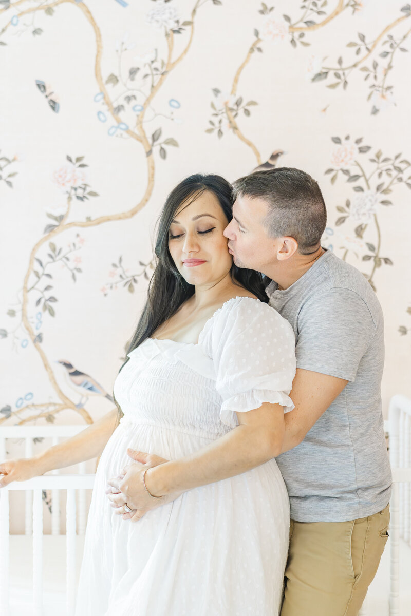 A couple stands in a pink nursery in front of a white crib in their Austin home. The husband stands behind his pregnant wife while wrapping his arms around her belly and kissing her on the cheek. The mother, dressed in a long white dress, closes her eyes.