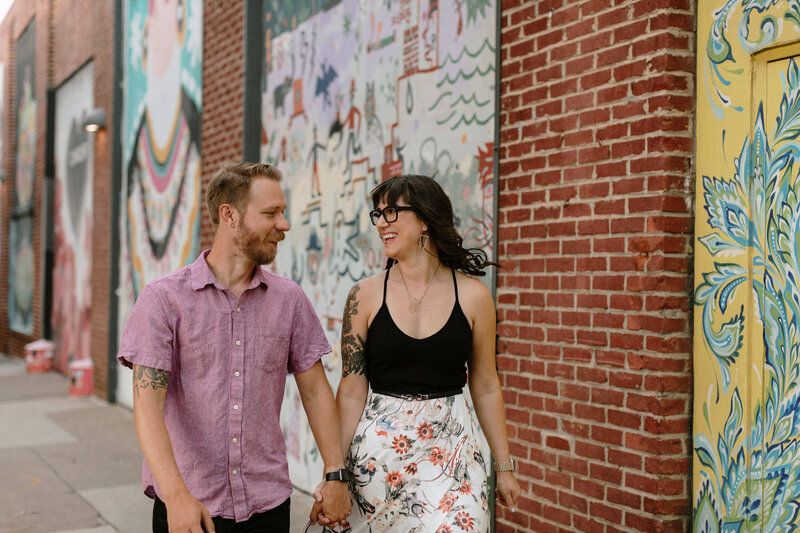 A couple walks hand in hand along a brick wall adorned with colorful murals, wearing stylish summer outfits.