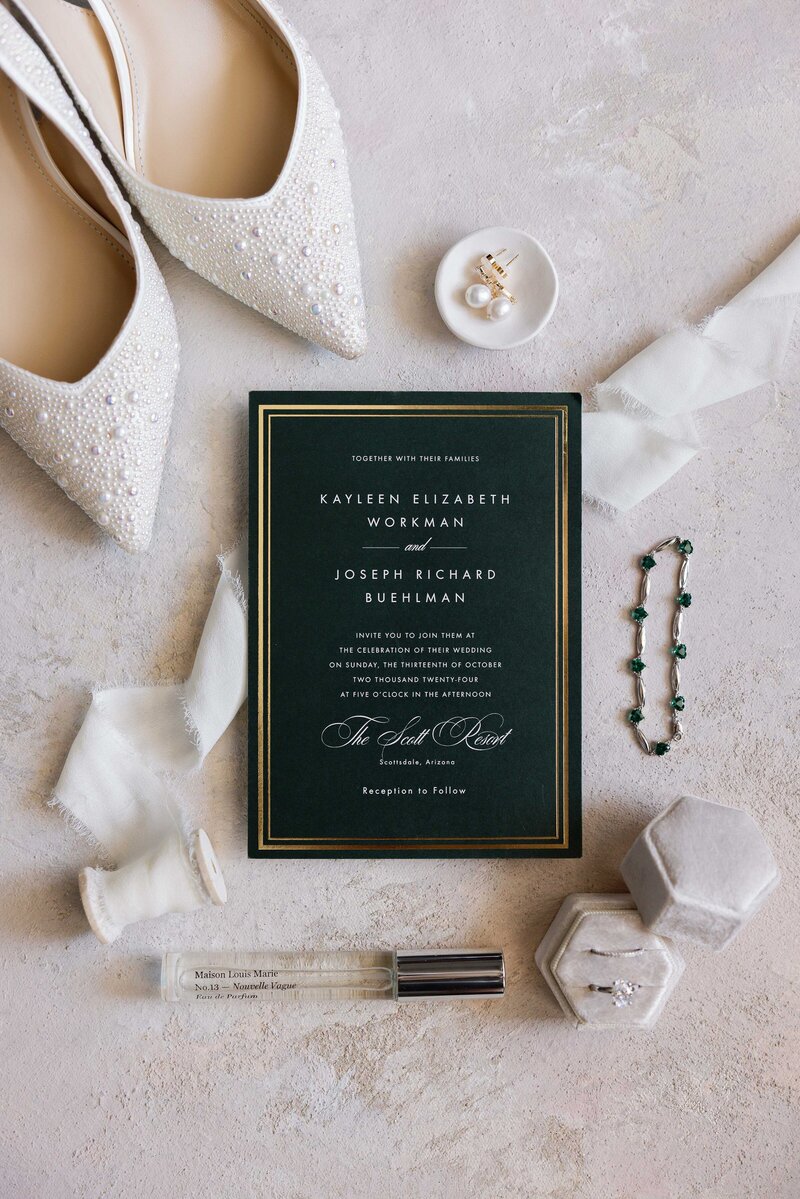 Dark green wedding invitation styled with bride’s jewelry, shoes, and silk ribbon.
