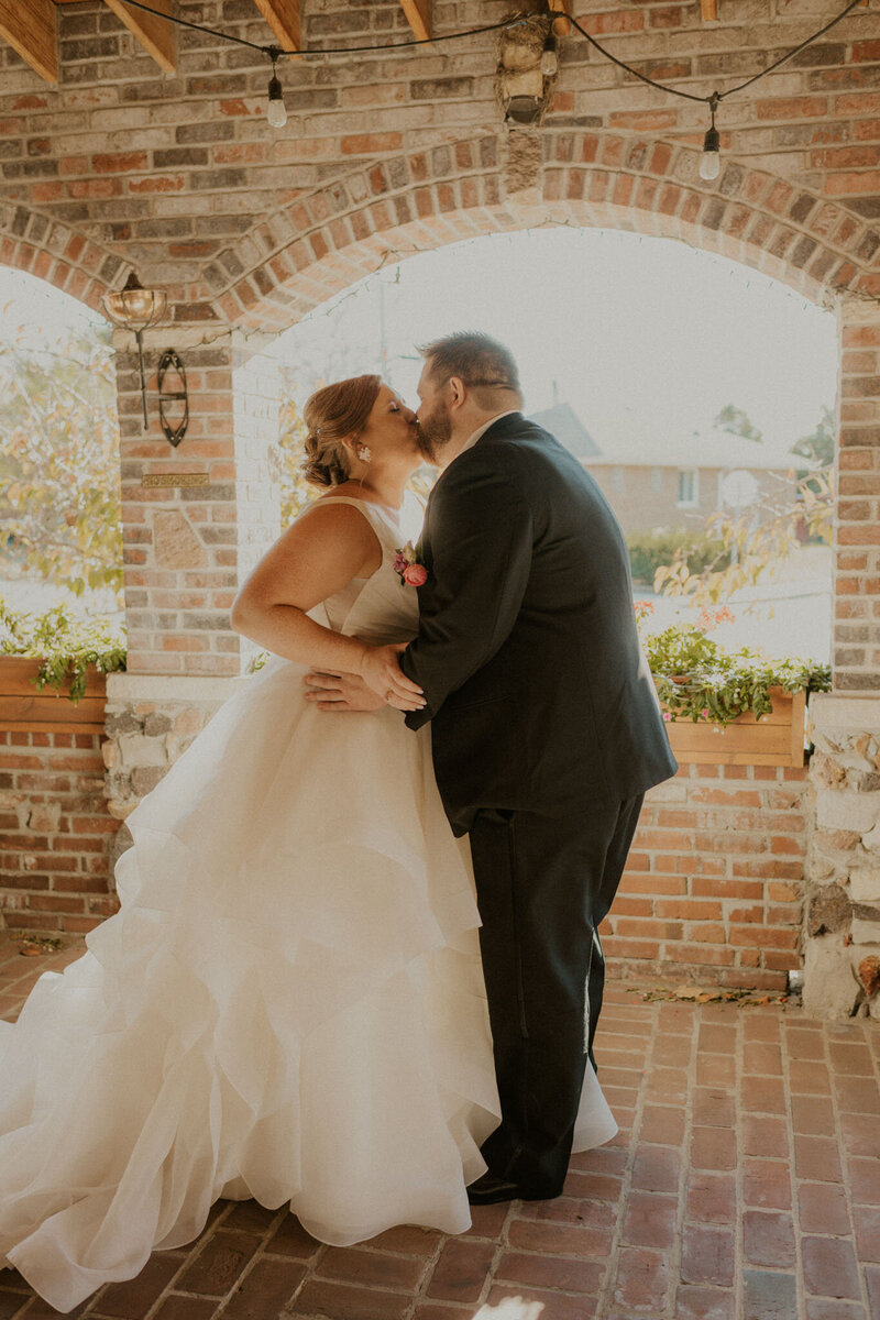 A bride and groom share a kiss under a brick archway on their wedding day, at Grbic Events, a wedding venue in St. Louis.
