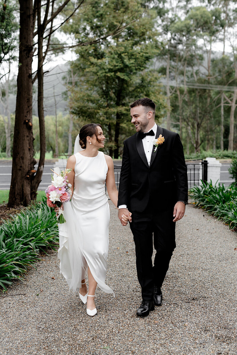 Bride and groom walking towards camera but looking at each other having a laugh. Bride wearing a silk dress with the train and veil over her arm. They are holding hangs also.