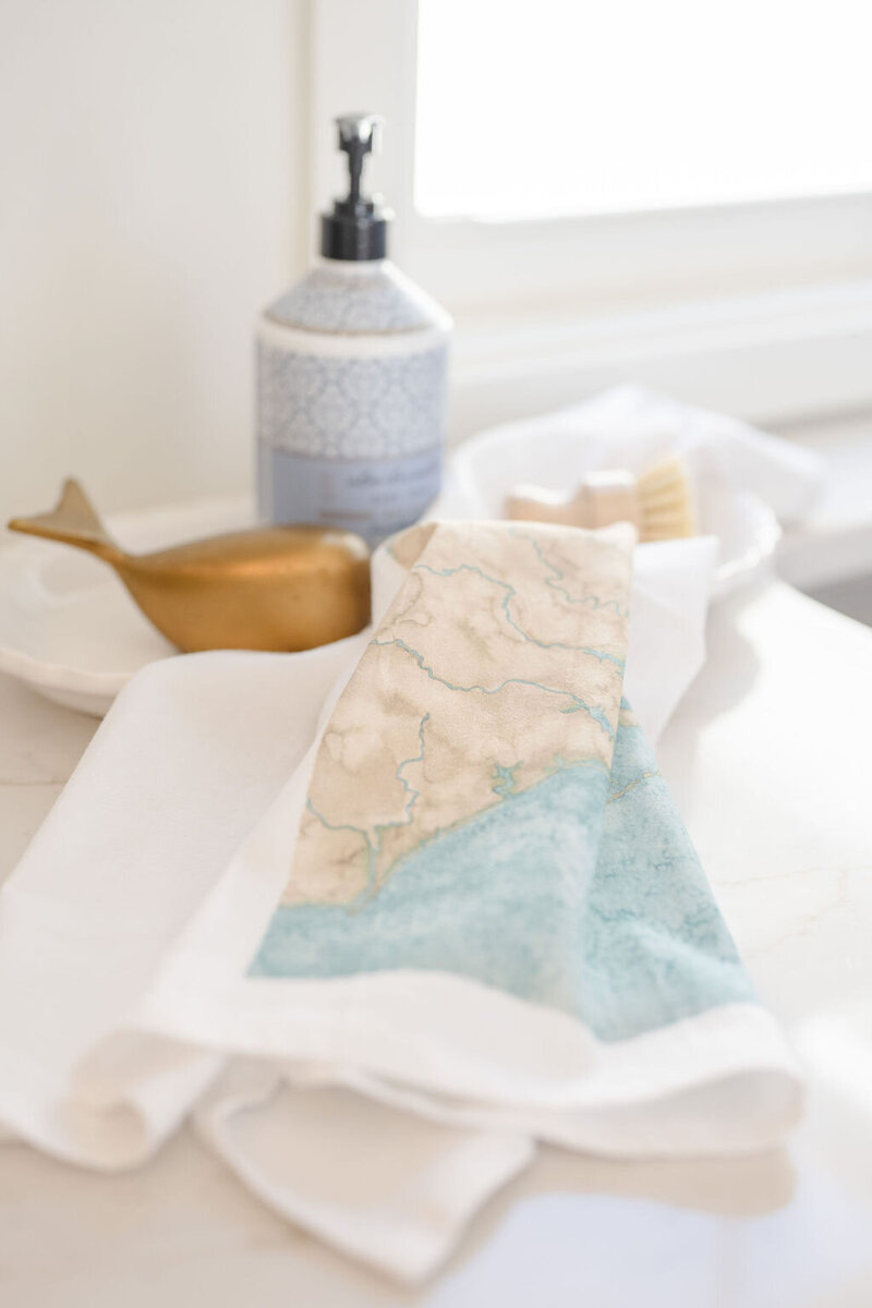 a tea towel with coastal watercolor artwork set over the counter with hand soap and accessories in the background in wilmington, north carolina