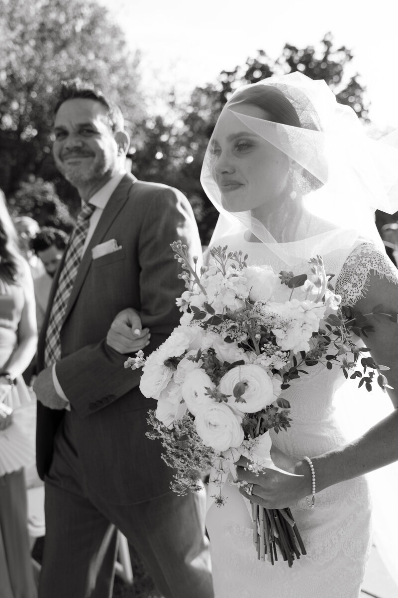 Black and white photo of bride walking down the aisle with her father, holding a bouquet of white flowers during outdoor ceremony.