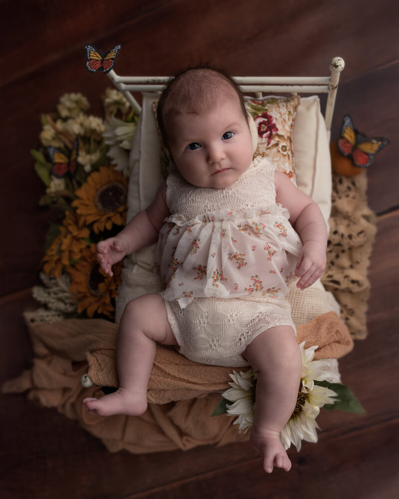  Baby girl milestone image with butterflies, floral textures, and a rustic vintage bed setting—captured in warm, earthy tones.