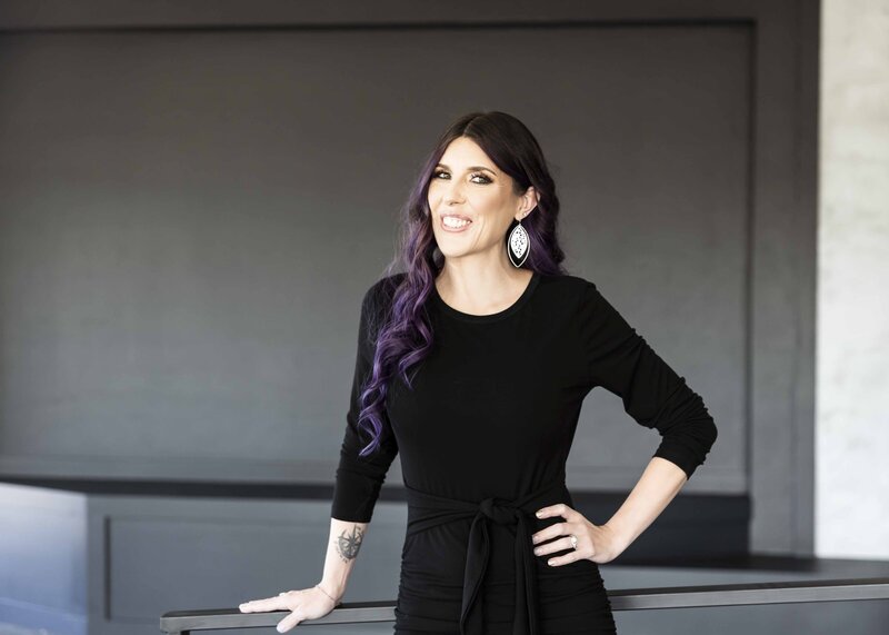Laura is wearing a black dress, and she smiles toward the camera with her left hand on her hip, and her right hand resting on a nearby railing. An open-circle tattoo is visible on this wrist. Laura has light skin, and her hair is long and purple with dark brown roots.