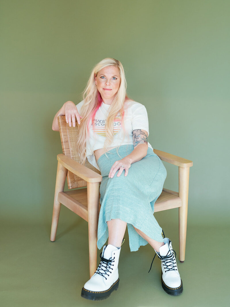 Kristen looking at the camera on a green background with a blue long skirt and white doc martens, a pink streak in her blonde hair