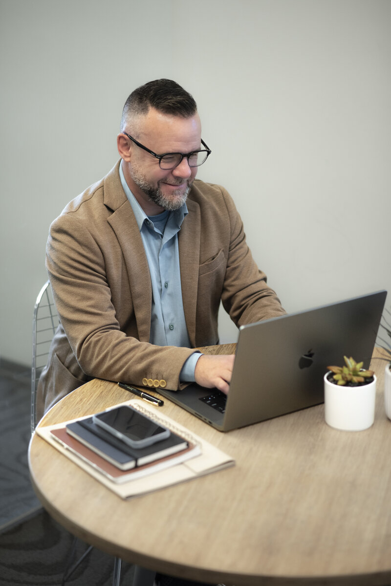 Matthew Kidwell in a brown blazer working on laptop and smiling - Financial Coach at Kidwell Coaching and Consulting 12
