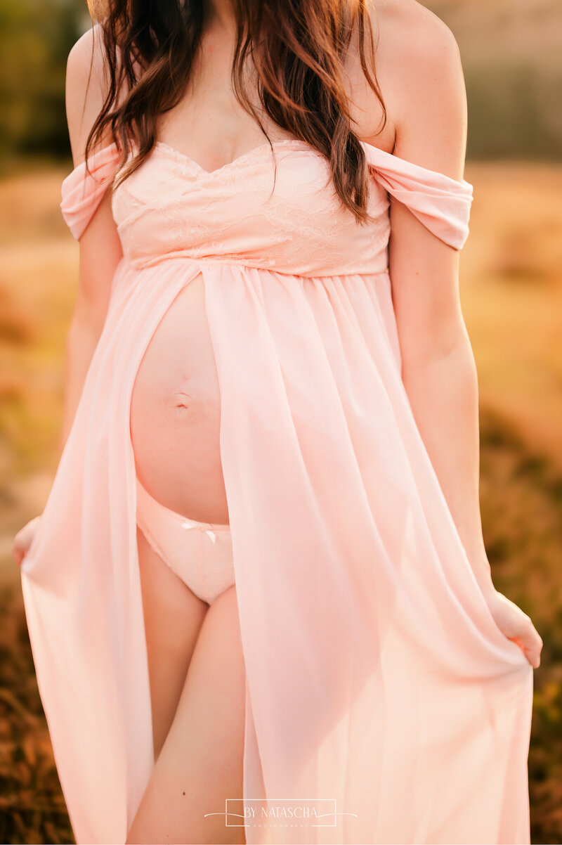 Foto van een zwangerschapsshoot met een roze jurk aan die open valt en de buik zichtbaar is door de openslaande jurk met plooien en bandjes van haar schouders af vallen voor een mooie sexy look