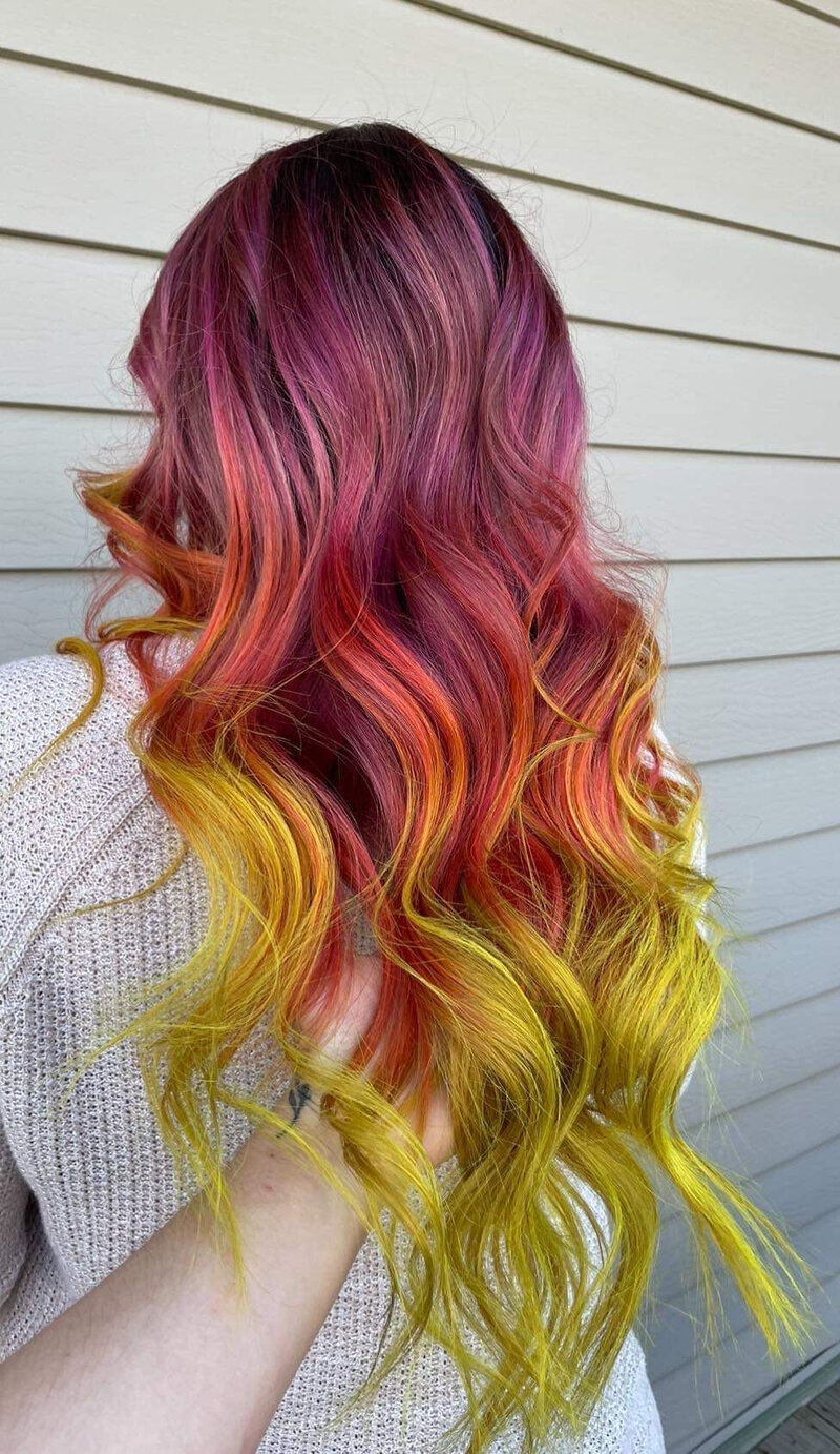 A girl with a curled purple, red, and yellow color melt on long hair, facing a tan building