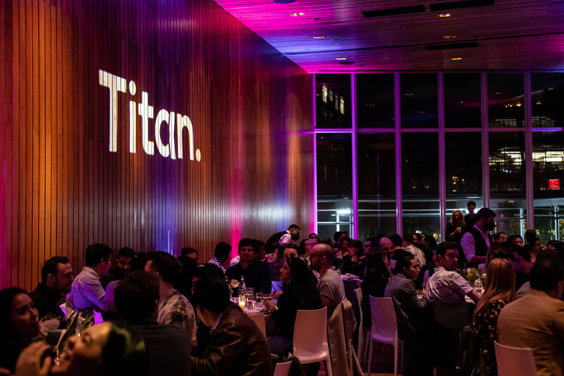 Guests seated at dinner tables during Titan Day 2 in New York, with the word “Titan.” projected in large white letters on a tall wooden wall illuminated by purple and orange lighting.