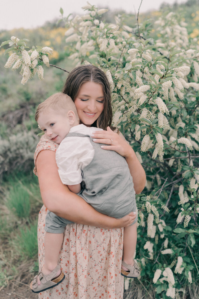 Joyful mom holding bump with wind in her hair, mountain ridges behind. CAPTURED BY Newborn Photography Salt Lake City, Newborn Photographer Riverton, Maternity Photographer Salt Lake City.