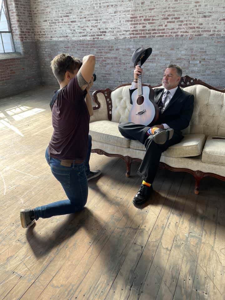 A photographer taking a picture of a person sitting on a couch holding a guitar 