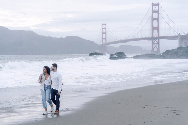 Engaged couple walking at baker beach with the golden gate bridge in the background