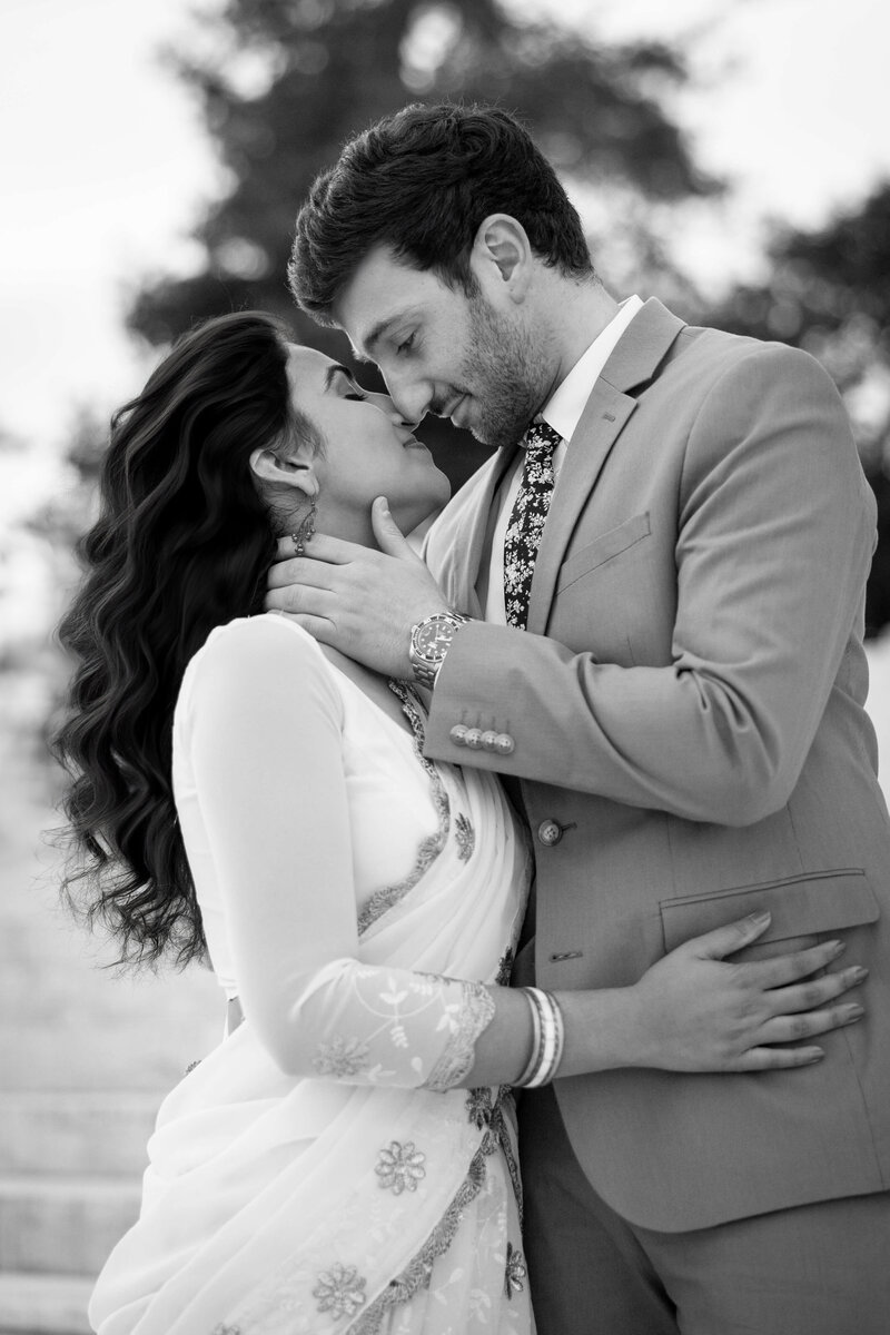 A couple in formal attire shares an intimate moment, gazing into each others eyes. The woman wears a traditional sari and bangles, while the man wears a suit with a floral tie. The background is softly blurred. Black and white image.