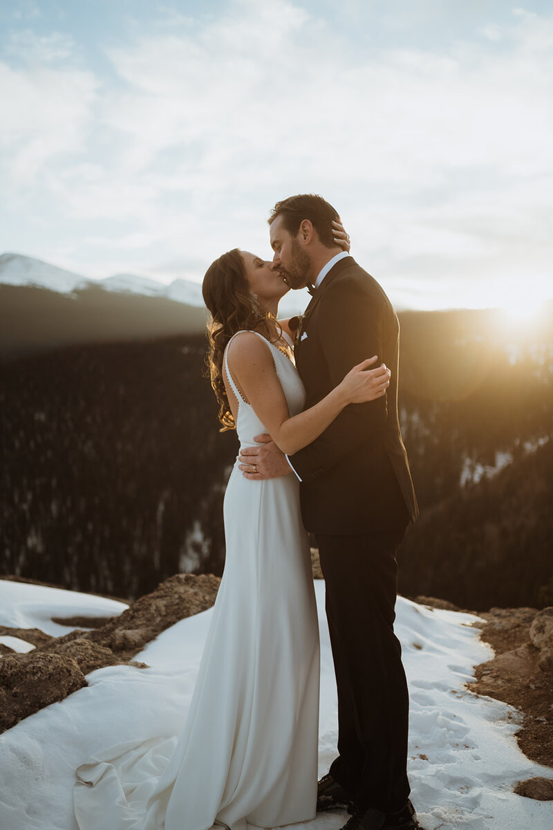 bride and groom kissing with the sun in the background. the mountains behind them are covered in snow, and the bride has her hand on the back of the grooms neck.