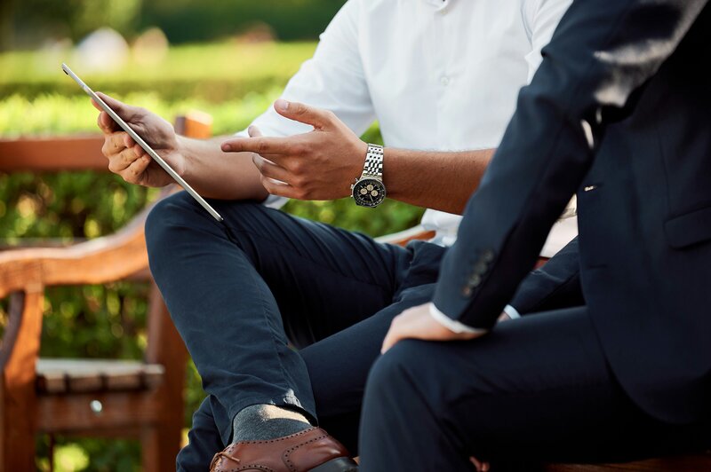 Two professionals sitting on an outdoor bench, one holding a tablet and pointing while having a discussion.