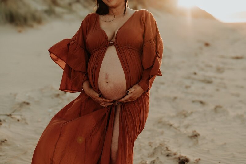 Close up shot of a pregnant woman's belly for a maternity session. She is wearing a rust colored dress that opens at her belly. The sunrises behind her