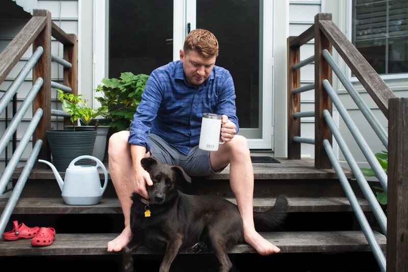 A man sits on the stairs outside his house while drinking coffee and petting a dog