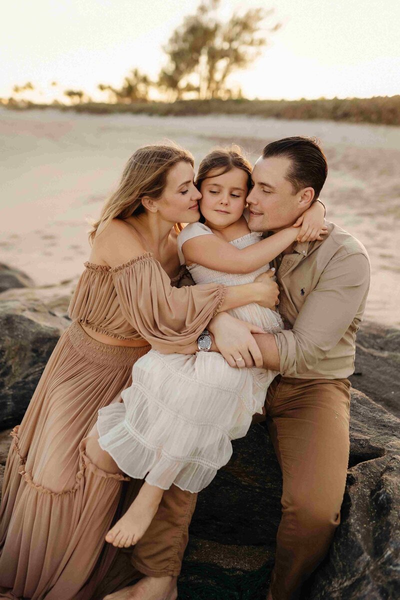Beach portrait of parents embracing their daughter on rocks at sunset, gentle expressions, warm light, ocean in the backgroun